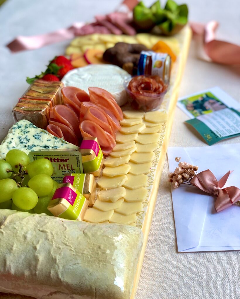 Uma tábua de madeira rústica retangular, servindo como base para uma variedade de petiscos gourmet. A tábua está repleta de cores vibrantes e texturas convidativas, criando uma composição visualmente atraente. Entre os petiscos, destacam-se: Queijos: Queijo brie, queijo faixa azul, queijo do reino em fatias finas, queijo provolone defumado em tiras. Charcutaria: Lombo canadense, salame italiano em fatias finas, copa em fatias finas. Frutas: Uvas verdes, morangos frescos, damasco desidratado. Detalhes da imagem: Tema: Tábua de frios gourmet, petiscos, aperitivos, gastronomia, festa, evento, recepção. Elementos: Tábua de madeira, queijos variados, frutas frescas, azeitonas, palitos de grissini. Emoção: Sofisticação, requinte, elegância, sabor, prazer.