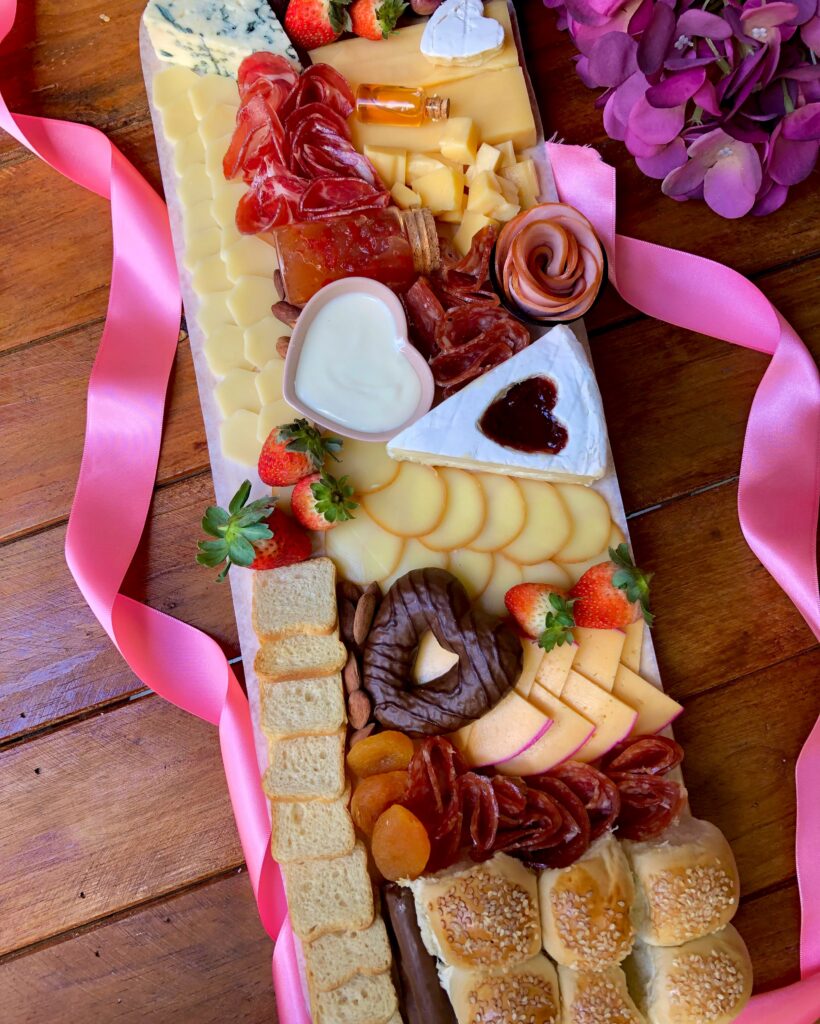 Uma tábua de madeira rústica retangular, servindo como base para uma variedade de petiscos gourmet. A tábua está repleta de cores vibrantes e texturas convidativas, criando uma composição visualmente atraente. Entre os petiscos, destacam-se: Queijos: Queijo brie, queijo faixa azul, queijo do reino em fatias finas, queijo provolone defumado em tiras. Charcutaria: Lombo canadense, salame italiano em fatias finas, copa em fatias finas. Frutas: Uvas verdes, morangos frescos, damasco desidratado. Detalhes da imagem: Tema: Tábua de frios gourmet, petiscos, aperitivos, gastronomia, festa, evento, recepção. Elementos: Tábua de madeira, queijos variados, frutas frescas, azeitonas, palitos de grissini. Emoção: Sofisticação, requinte, elegância, sabor, prazer.